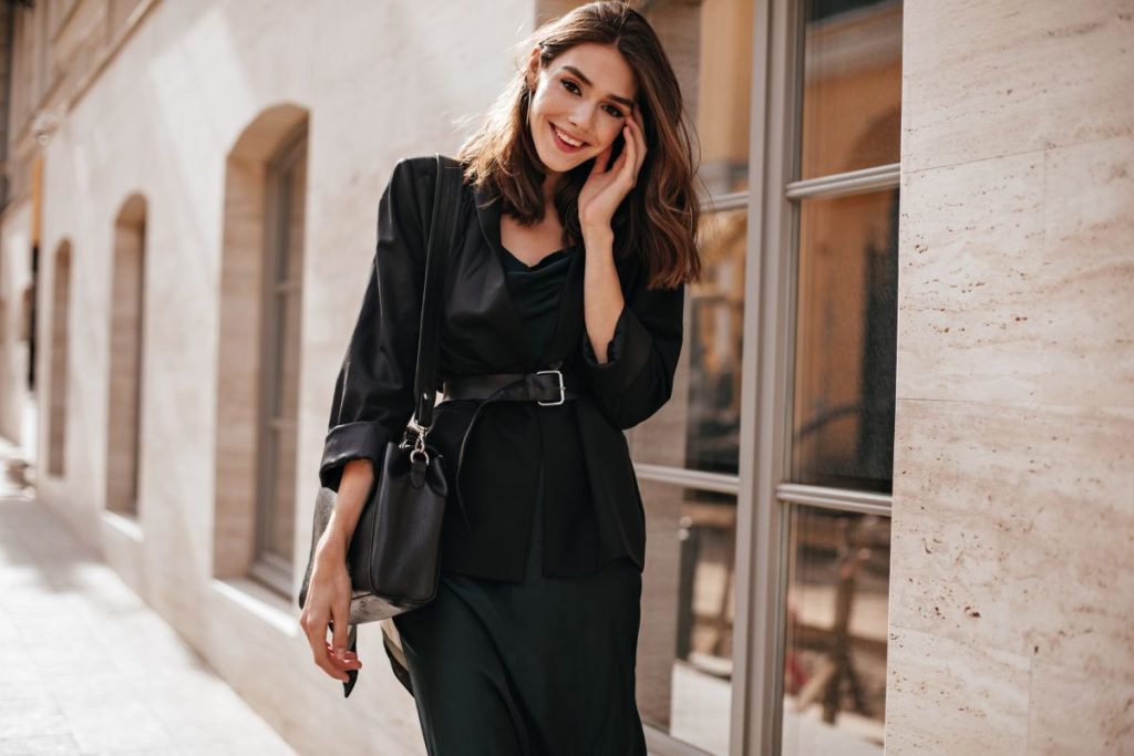 Cute slim girl with brown wavy hairstyle, bright makeup, dark outfit of long dress, oversized jacket, belt on waist and trendy bag, standing on daylight street in city and smiling