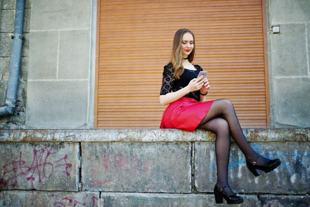Portrait of girl with black choker on her neck, red leather skirt and mobile phone at hand against orange shutter.