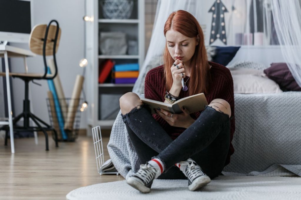 Redhead girl thinking over a notebook while sitting on a floor in her room
