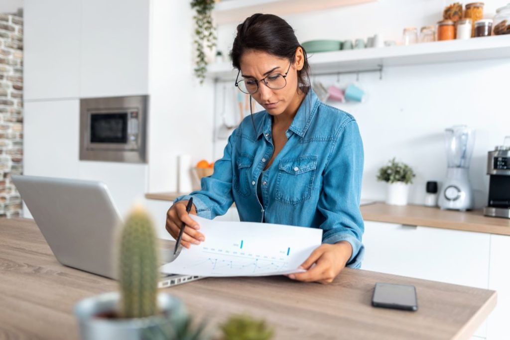 Shot of pretty young woman reviewing paperwork and working on a laptop in the kitchen at home.