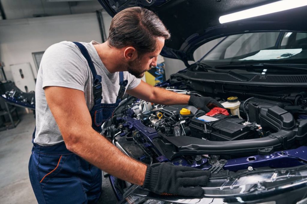 Man auto technician looking carefully inside car engine and touching its battery with hand during examination