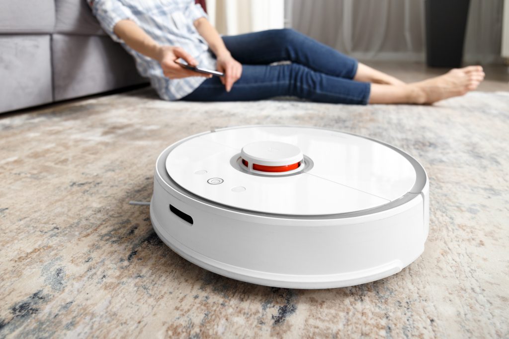 A girl sits on the floor in the living room and controls a robotic vacuum cleaner using a smartphone. Smart House. The concept of smart home appliances, housework and technology.