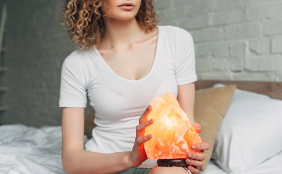 cropped view of curly woman in homewear holding Himalayan salt lamp in bedroom