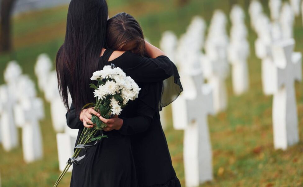 Embracing each other and crying. Two young women in black clothes visiting cemetery with many white crosses. Conception of funeral and Close Friend death